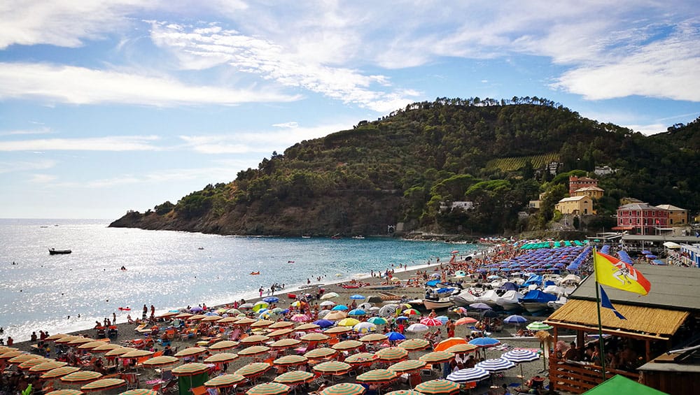 A spacious beach in Bonassola, Liguria. The shore is full of colorful sun umbrellas, with people swimming and sunbathing. In the background, the ligurian terracings and vineyards can be seen.
