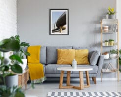 A black framed print of Ponte del Mare in Pescara, Italy hanging in a room with a grey an yellow sofa, with many plants and wooden decor. By Photographer Scott Allen Wilson.