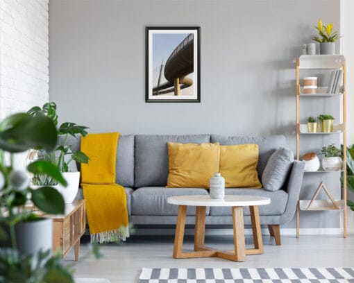 A black framed print of Ponte del Mare in Pescara, Italy hanging in a room with a grey an yellow sofa, with many plants and wooden decor. By Photographer Scott Allen Wilson.