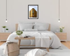 A black framed print of a symmetric yellow house in Pescara, Italy, hanging in a minimal bedroom with white and wooden decor. By Photographer Scott Allen Wilson.