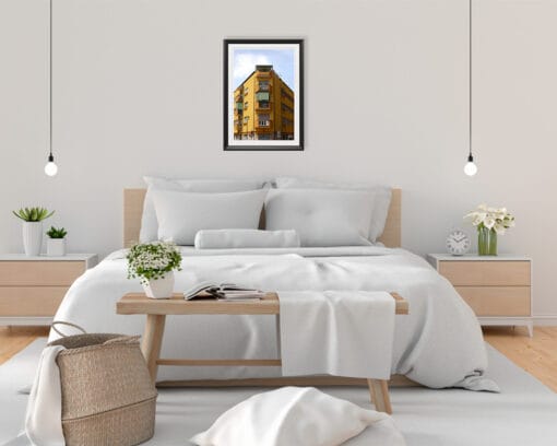 A black framed print of a symmetric yellow house in Pescara, Italy, hanging in a minimal bedroom with white and wooden decor. By Photographer Scott Allen Wilson.