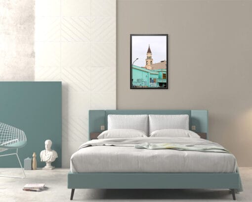 A print of a clock tower emerging from the back of a bright turquoise building covered in graffiti in Pescara, Italy, hangs in a minimal bedroom with a blue bed and furniture. By Photographer Scott Allen Wilson.