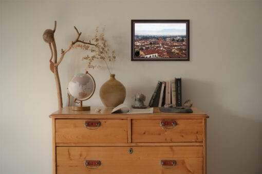 A dark brown framed print of a view of Lucca, Italy, hanging in a boho room with wooden decor. By Photographer Scott Allen Wilson.