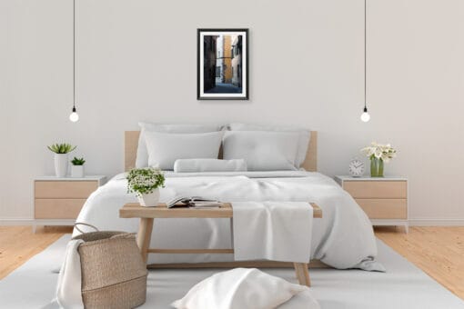 A black framed print of a view of Via Del Toro in Lucca, Italy, hanging in a minimal bedroom with wooden decor. By Photographer Scott Allen Wilson.