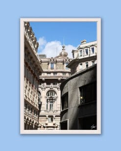 Wooden framed artistic print of a Genovese street scene, where grand adorned structures stand behind a curved, modern one and under a bright blue sky.