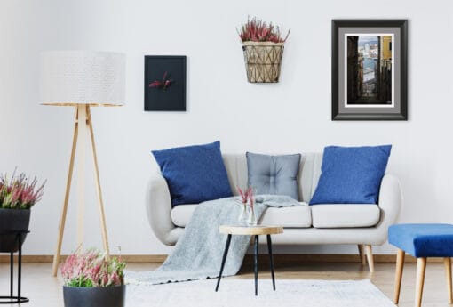 A brightly lit living room accentuates the glimmer of light between the shaded houses of the image, offering more depth to the white room’s walls. Navy blue pillows lay on the couch, trying to imitate the colors of the sea, while vivid plants donate a touch of color to the scene.