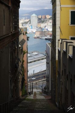 A narrow street in Genova, Italy, descends to the port of the city, while in the distance a beautiful cityscape opens up among the tall walls. Photo captured by Travel Photographer Scott Allen Wilson.