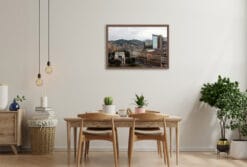 The warm tones of this shot harmonize beautifully with the cozy, earthy decor of this minimalist kitchen. The framed cityscape adds a sophisticated touch to the room, balancing the natural elements of plants and soft wood with a sense of urban and historical elegance.