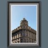 A black wooden framed piece of art of a gorgeous historical construction, filled with windows and decorative details, stands proud beneath a serene blue backdrop.